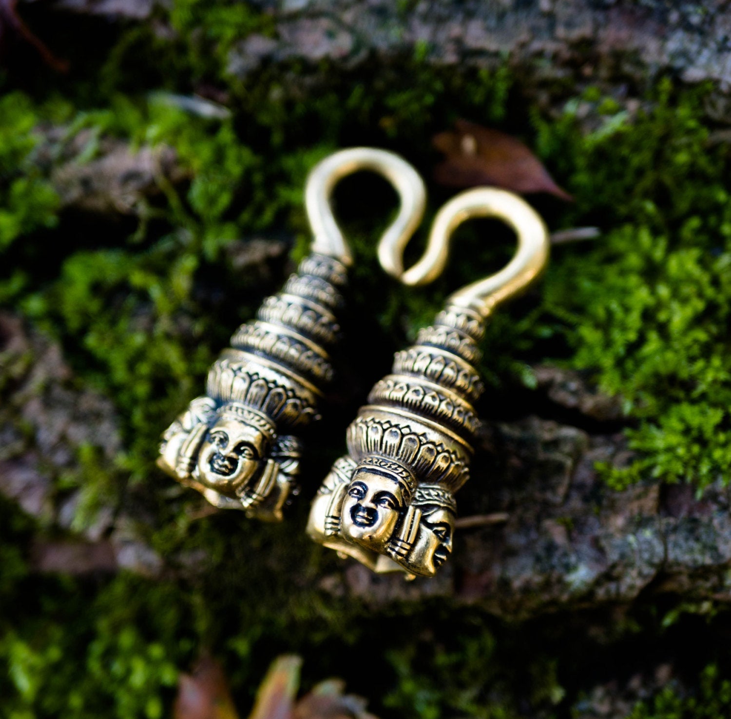 Two intricately designed ANGKOR WAT Heavy Ear Weights in Gold from Kaya Movement lie side by side on a moss-covered surface. Each 6 gauge piece features a detailed Buddha carving at the bottom, with ornate patterns leading up to a curved hook at the top. The background consists of green moss and textured bark.